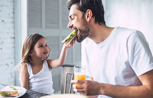 Niña convidando a su padre con un sandwich - Un padre le lee a sus hijos dentro de una carpa en el living de una casa - ¿Qué es la paternidad activa?